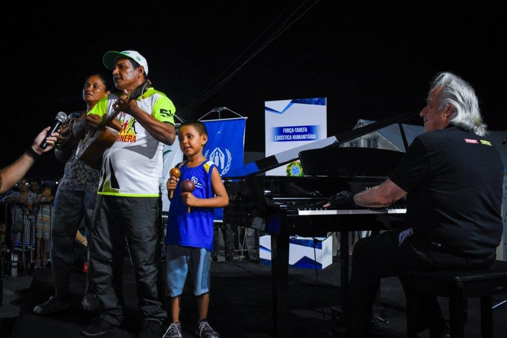 Guillermo, Katty e o pequeno Arnel se apresentando no palco ao lado do maestro brasileiro João Carlos Martins, enquanto a população do abrigo cantava junto as músicas tocadas