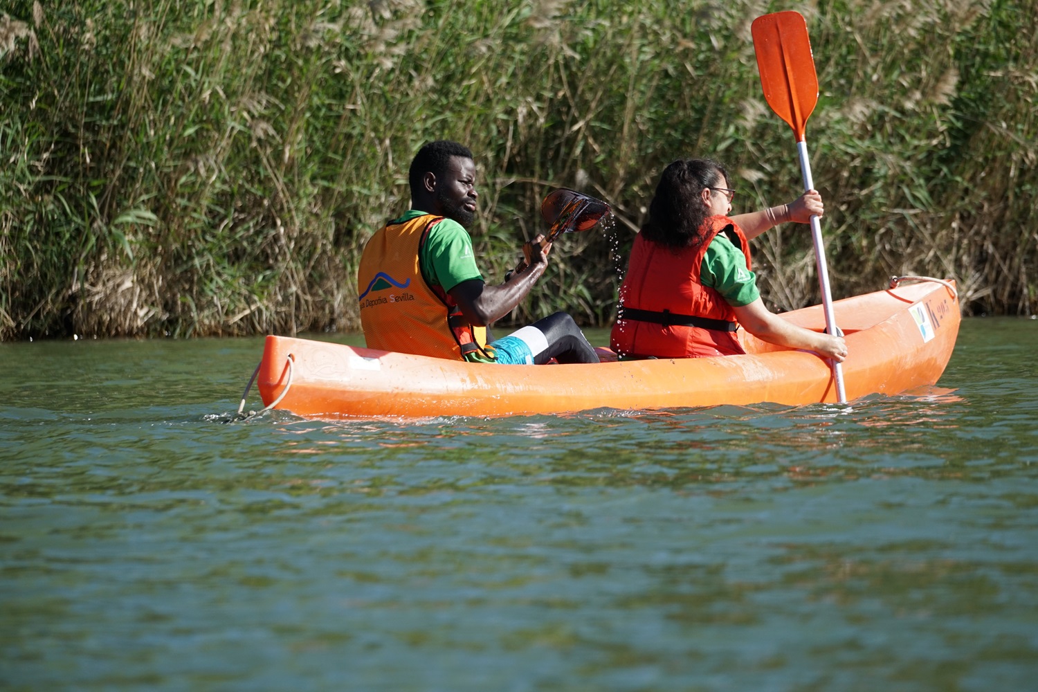 Aliou y una compañera de kayak reman en el Gualquivir junto a la orilla cubierta de vegetación.