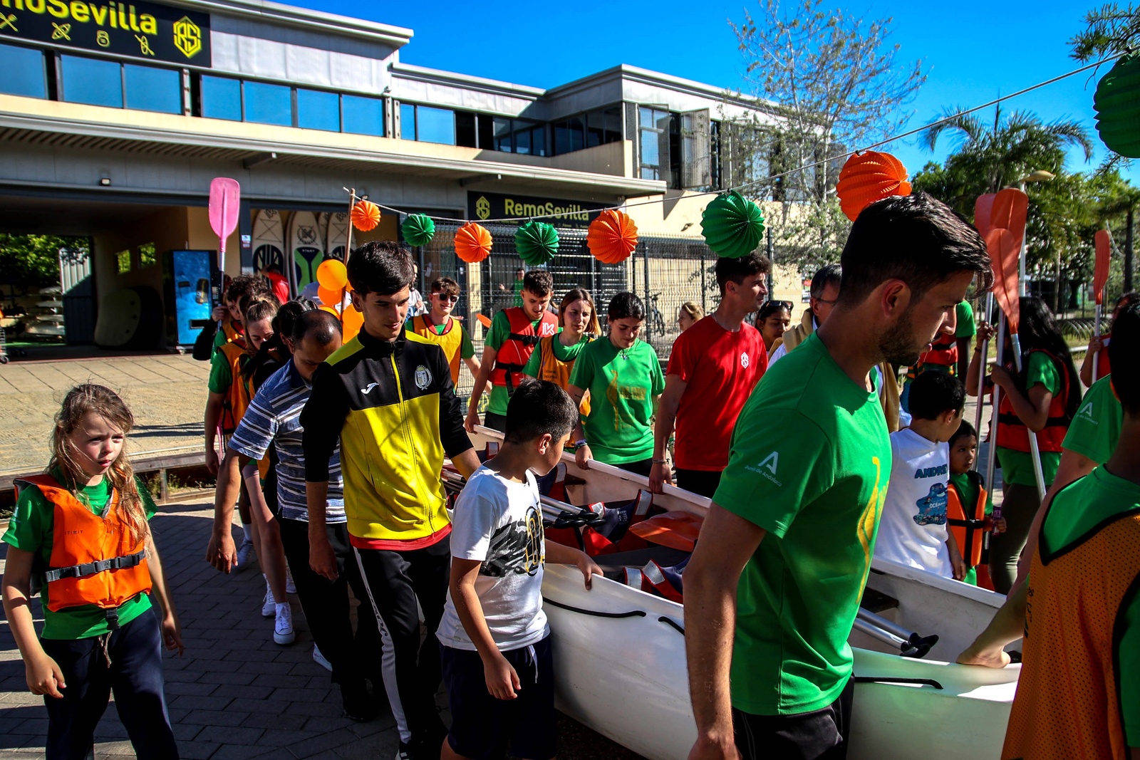 Varias personas de distintas edades transportan la canoa desde el Club de Remo Sevilla hacia el Guadalquivir.