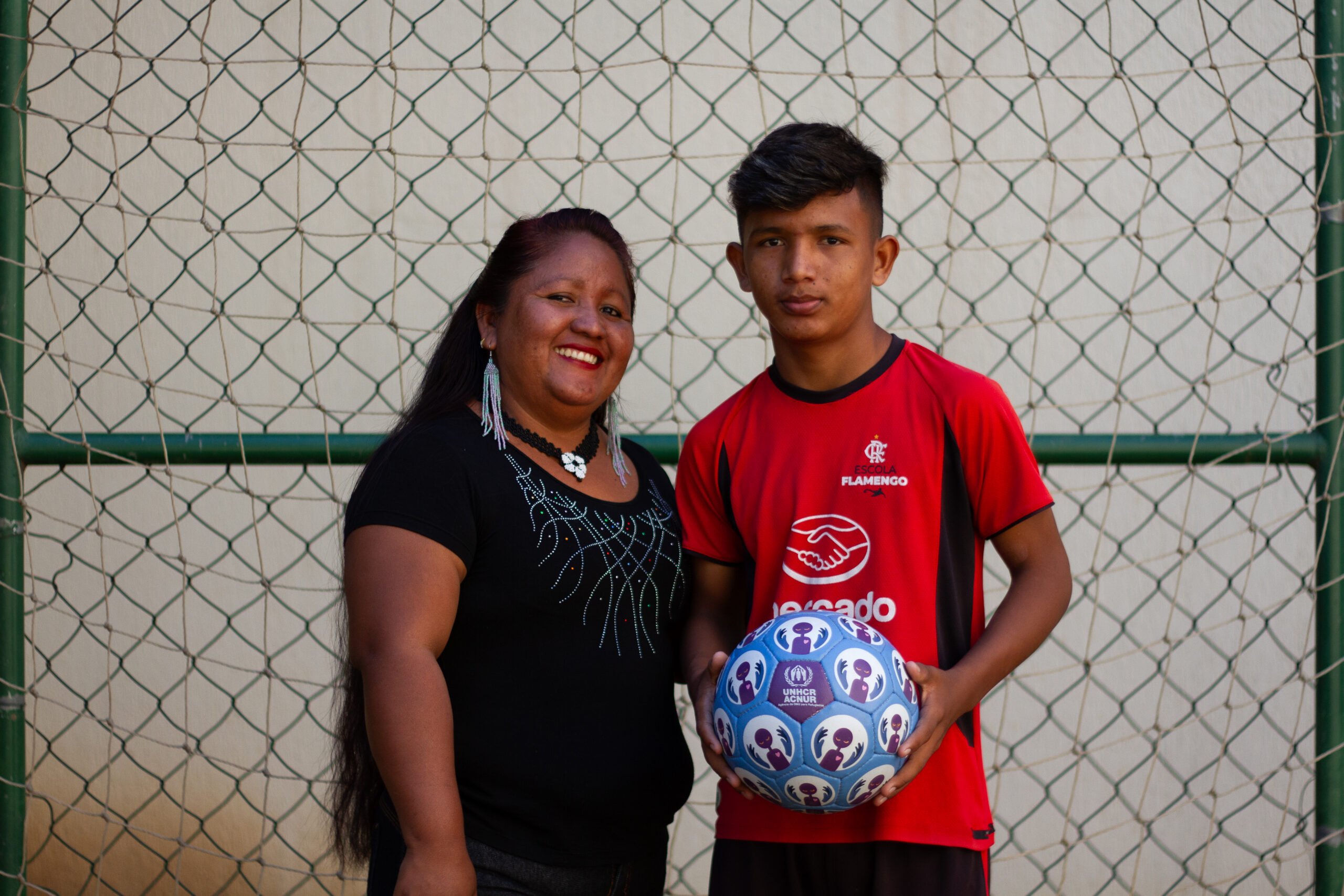 Do lado esquerdo, Josefina Moraleda, indígena warao venezuelana usando uma camisa preta e brincos grandes, e seu filho Greyver Moraleda, de 13 anos, vestindo o uniforme da Escola do Flamengo, com uma bola de futebol do ACNUR azul, roxa e branca.
